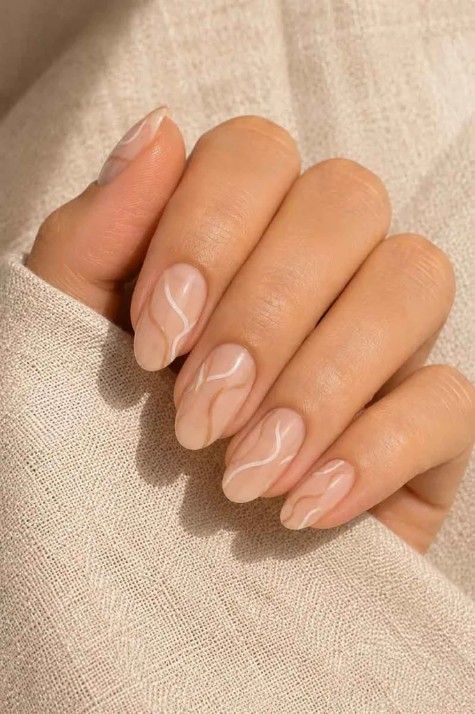 Close-up portrait of a hand with medium oval nails featuring loose freehand line nail art in warm white and tan tones on a nude base resting on textured cream linen fabric under soft side lighting.