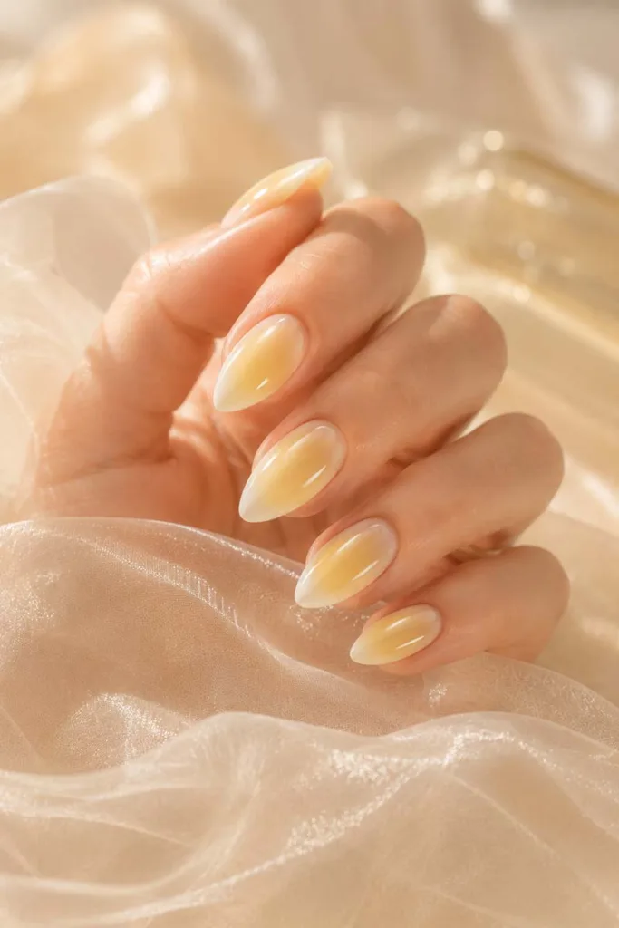 Dreamy close-up portrait of butter yellow aura nails fading into milky nude edges with a glossy jelly finish above sheer translucent fabric in soft diffused sunlight.