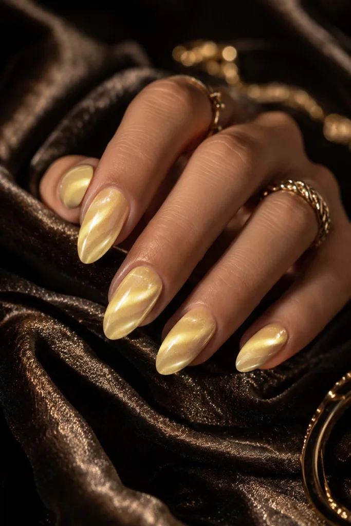 Dramatic close-up portrait of almond-shaped butter yellow velvet cat-eye nails with reflective magnetic shimmer against dark champagne velvet fabric and gold accessories.