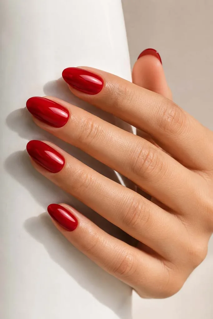 Close-up portrait of elegant almond-shaped nails painted in vivid glossy cherry red polish resting against a clean white ceramic surface under soft natural light.
