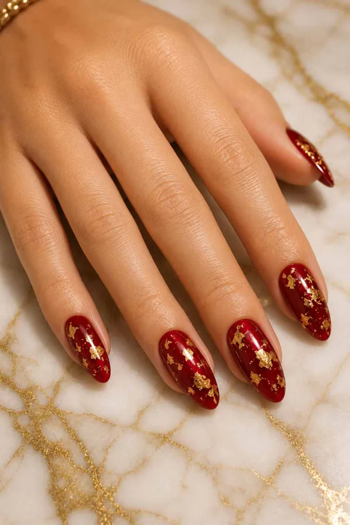 Close-up portrait of glossy deep red almond-shaped nails decorated with scattered gold foil flakes resting on a white marble surface with gold veining under warm studio lighting.