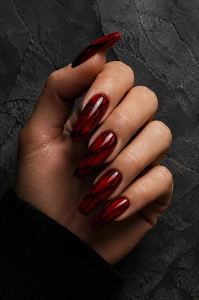 Close-up portrait of long coffin-shaped nails featuring deep red polish with black marble veining on a glossy finish against a dark charcoal stone background with moody lighting.