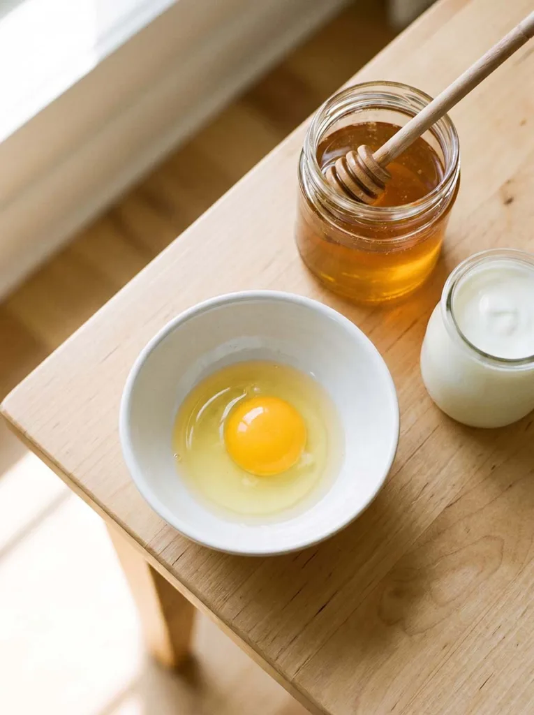 A top-down shot of a small white ceramic bowl with a freshly cracked egg inside. A jar of raw honey with a wooden honey dipper and a small pot of plain yogurt placed beside it on a light wooden surface. Soft natural daylight from the side. Warm tones, clean and simple arrangement.