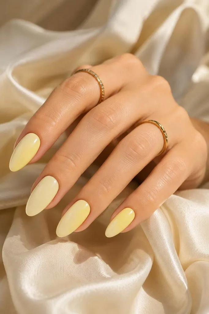 Close-up portrait of glossy butter yellow almond nails with a creamy pastel finish resting on ivory silk fabric with delicate gold rings and soft luxury lighting.