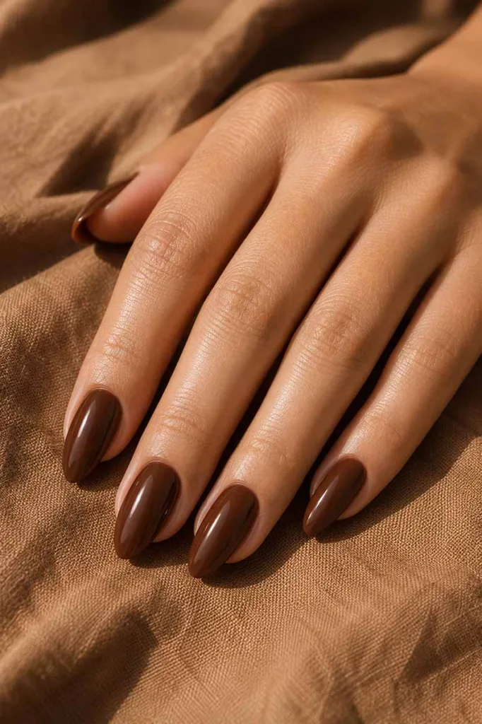 Close-up portrait of a hand with medium almond nails painted in a deep warm brown glossy polish resting on caramel-toned linen fabric with soft side lighting creating rich editorial shadows.