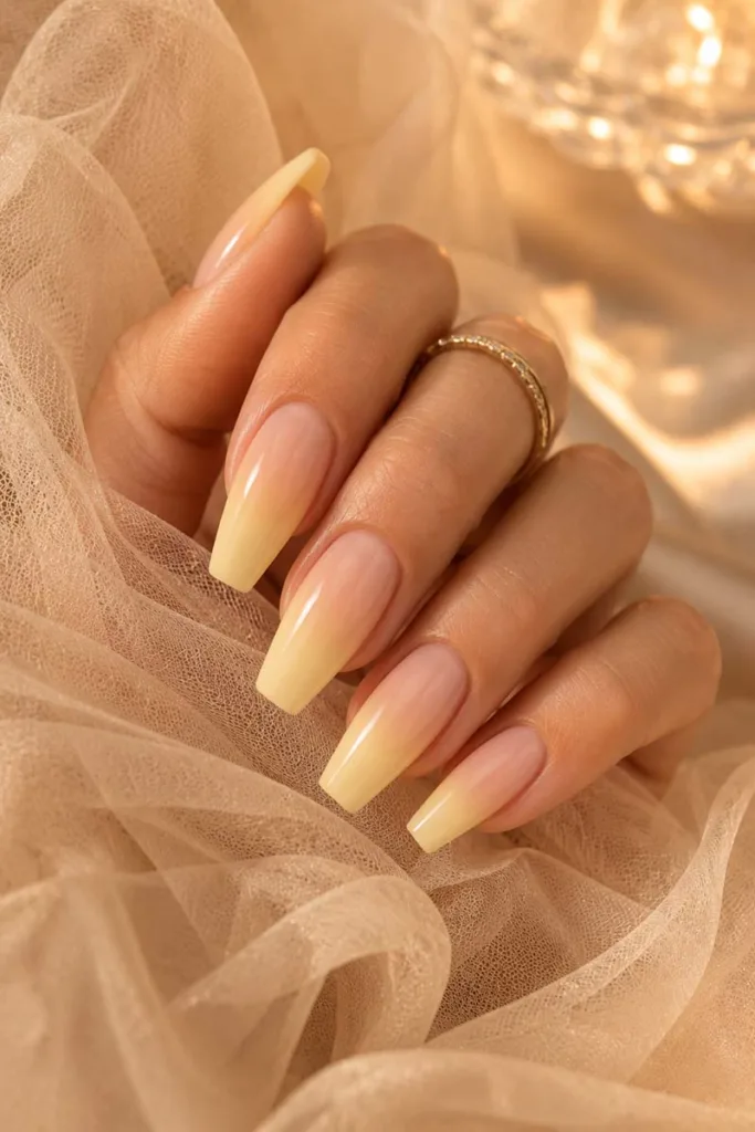 Elegant close-up portrait of glossy coffin nails with a seamless nude-to-butter-yellow ombre gradient resting on beige tulle fabric in warm champagne lighting.
