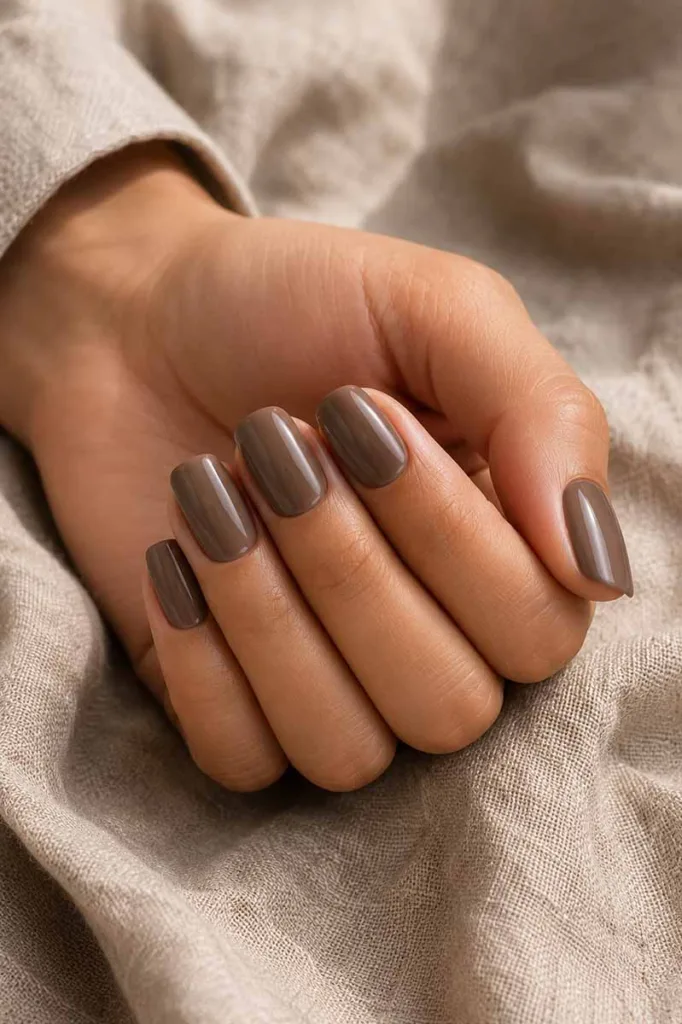 Close-up portrait of a hand with medium-length squoval nails painted in a deep warm taupe glossy polish resting on textured beige linen fabric under soft natural window light.
