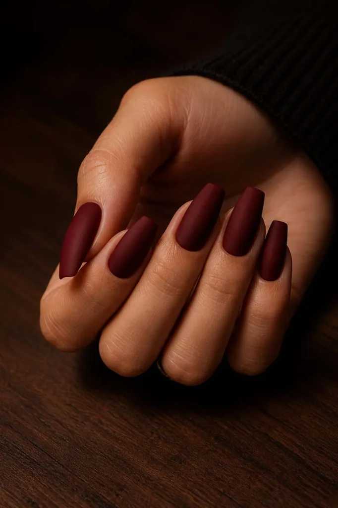Close-up portrait of medium coffin-shaped nails painted in deep matte burgundy red resting on a dark walnut wood surface under warm moody side lighting with soft dramatic shadows.