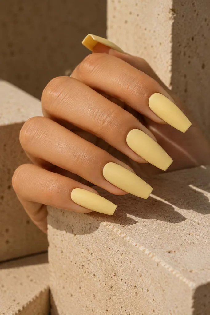 Close-up portrait of long coffin nails in matte pastel yellow polish against textured beige stone surfaces with warm editorial lighting and minimalist styling.