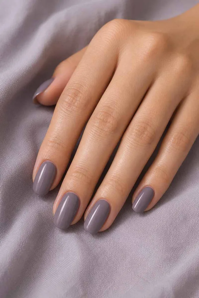 Close-up portrait of a hand with medium oval nails painted in a muted purple-grey polish resting on soft lilac-grey fabric under diffused natural light with cool neutral tones and a minimal editorial aesthetic.