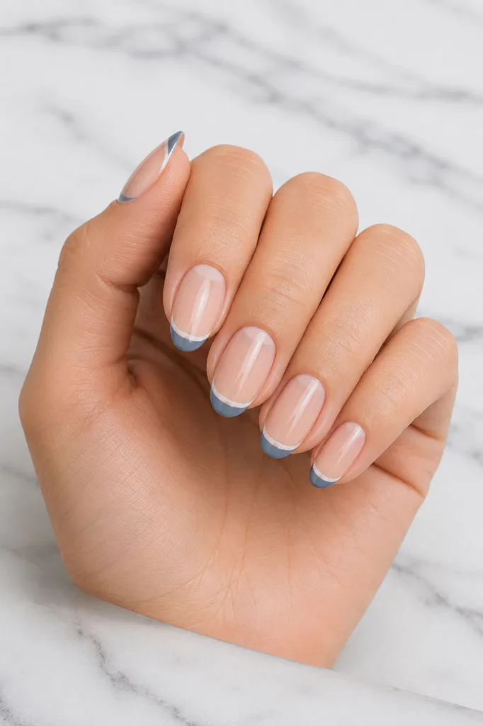 Close-up portrait of a hand with medium-length nude nails featuring thin colored French tips separated by a visible negative space gap against a cool white marble surface with soft grey veining.