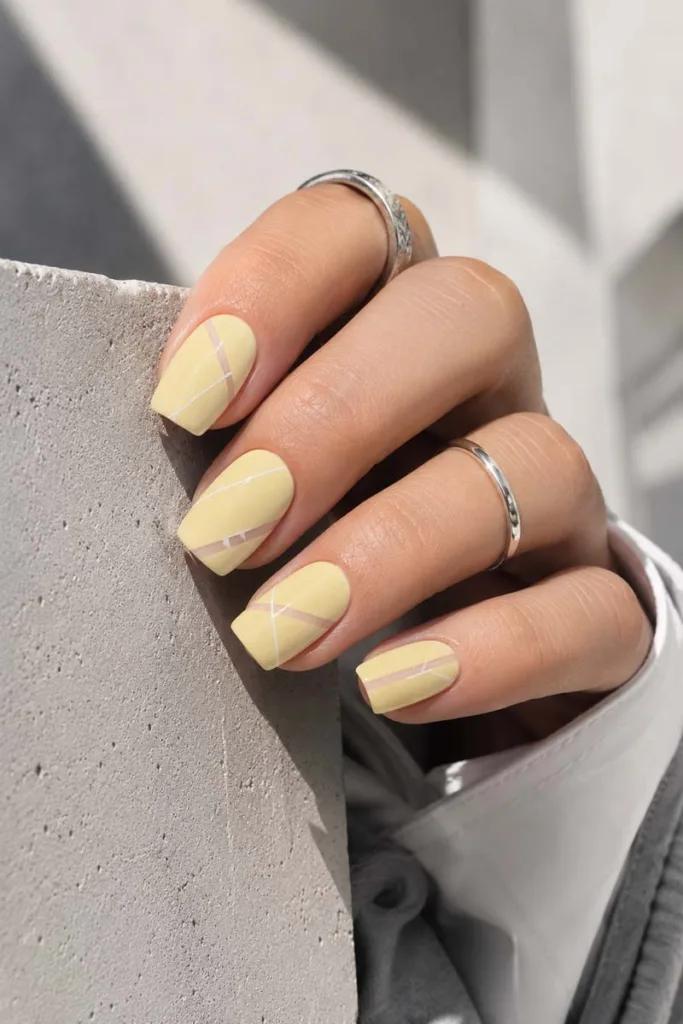 Modern close-up portrait of soft square pale butter yellow nails with thin white geometric lines and negative-space detailing against a textured concrete background.