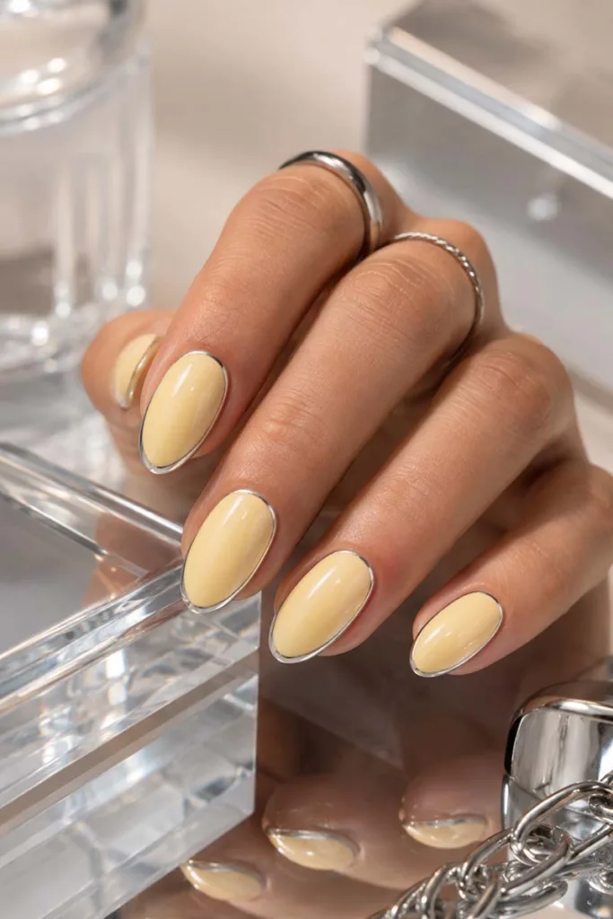 Sleek close-up portrait of creamy pastel yellow almond nails with thin silver chrome outlines resting on mirrored acrylic surfaces under bright studio lighting.