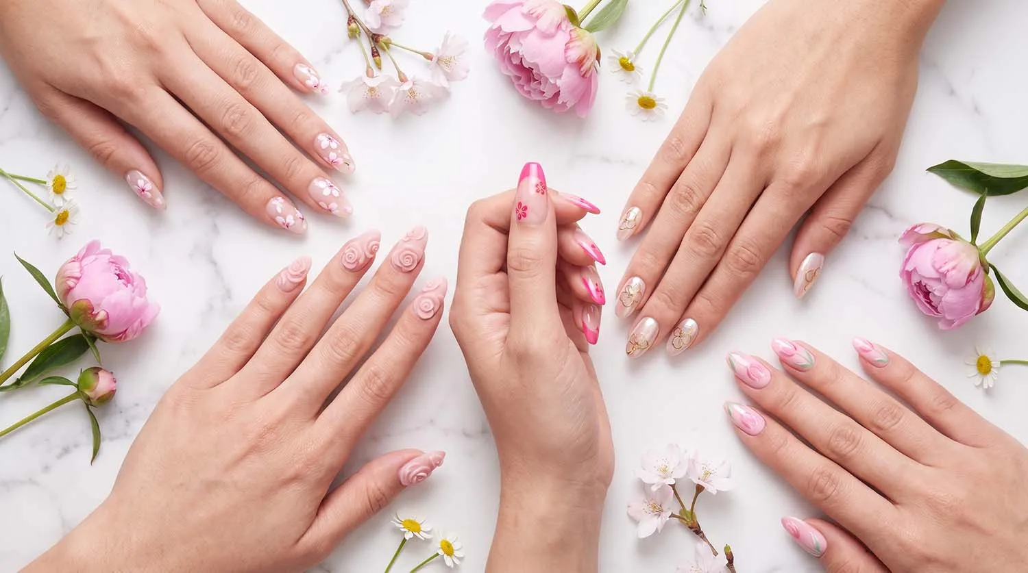 Five hands displaying different pink floral nails styles including milky cherry blossom sculpted 3D roses hot pink hibiscus French tips pearl chrome blossoms and blooming gel tulips on a white marble surface with fresh pink peonies and white daisies scattered around