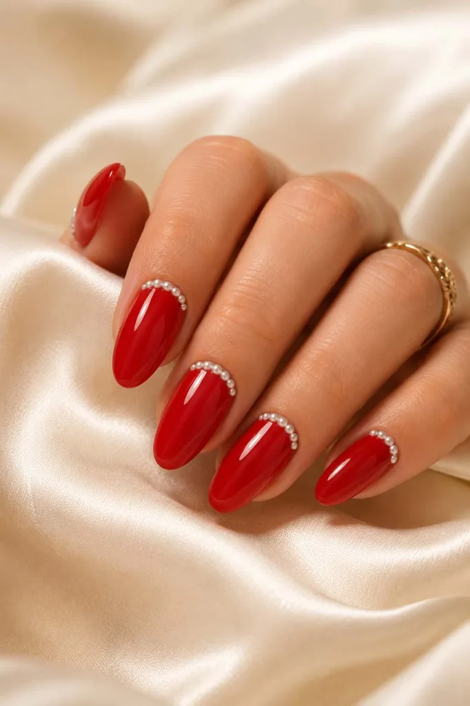 Close-up portrait of glossy scarlet red almond-shaped nails decorated with tiny white pearl beads along the cuticle line resting on cream satin fabric under warm diffused lighting.