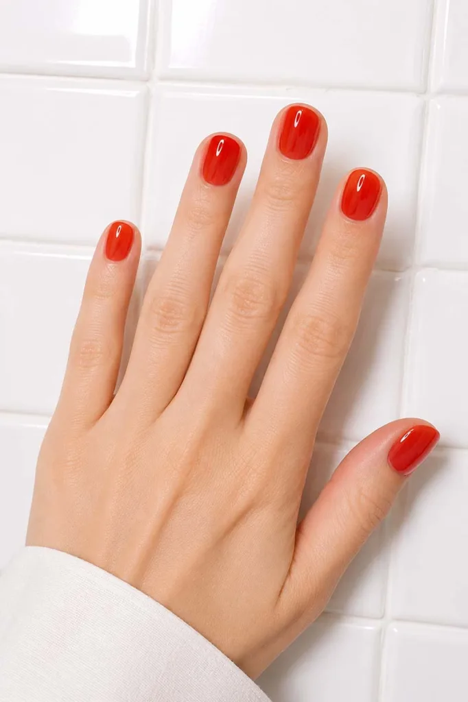 Close-up portrait of short round nails painted in semi-translucent tomato red jelly gel polish against a bright glossy white tiled surface with a glass-like glossy finish and youthful clean styling.
