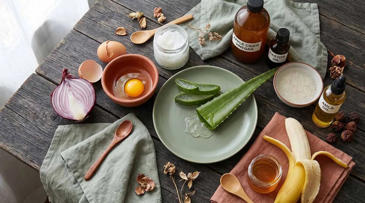 Nine natural remedies for weak hair at home including onion, egg, coconut oil, aloe vera, apple cider vinegar, castor oil, rice water, amla oil, and banana honey mask arranged on dark wood