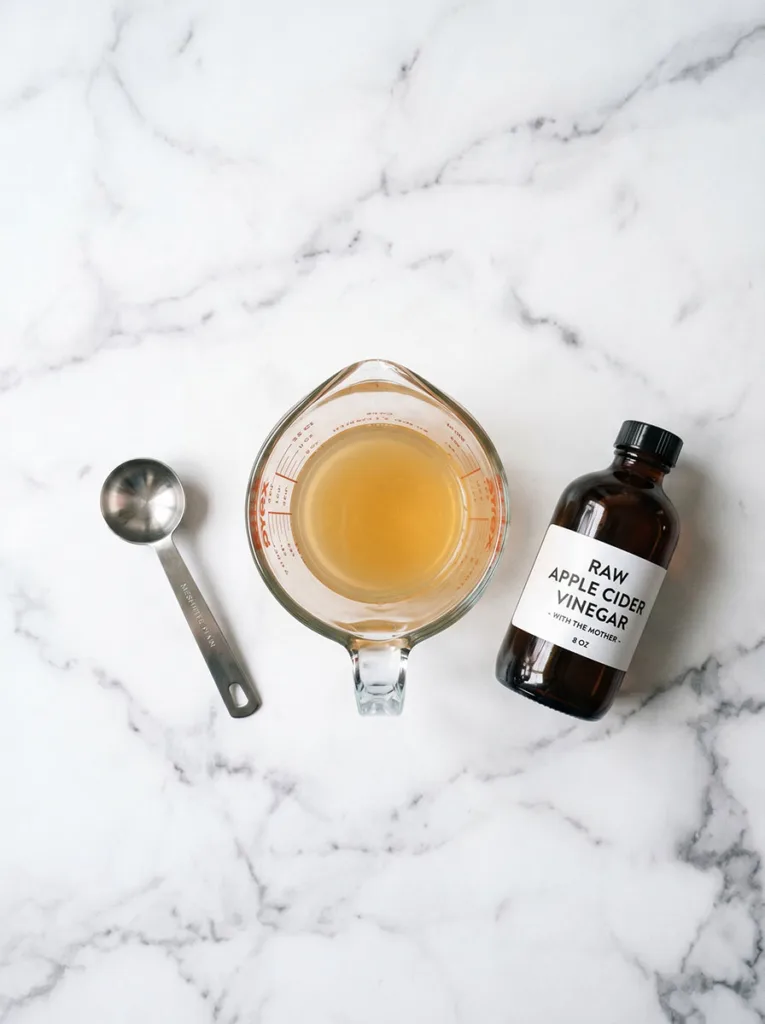 Top-down view of diluted apple cider vinegar in a measuring cup next to a dark glass bottle on white marble