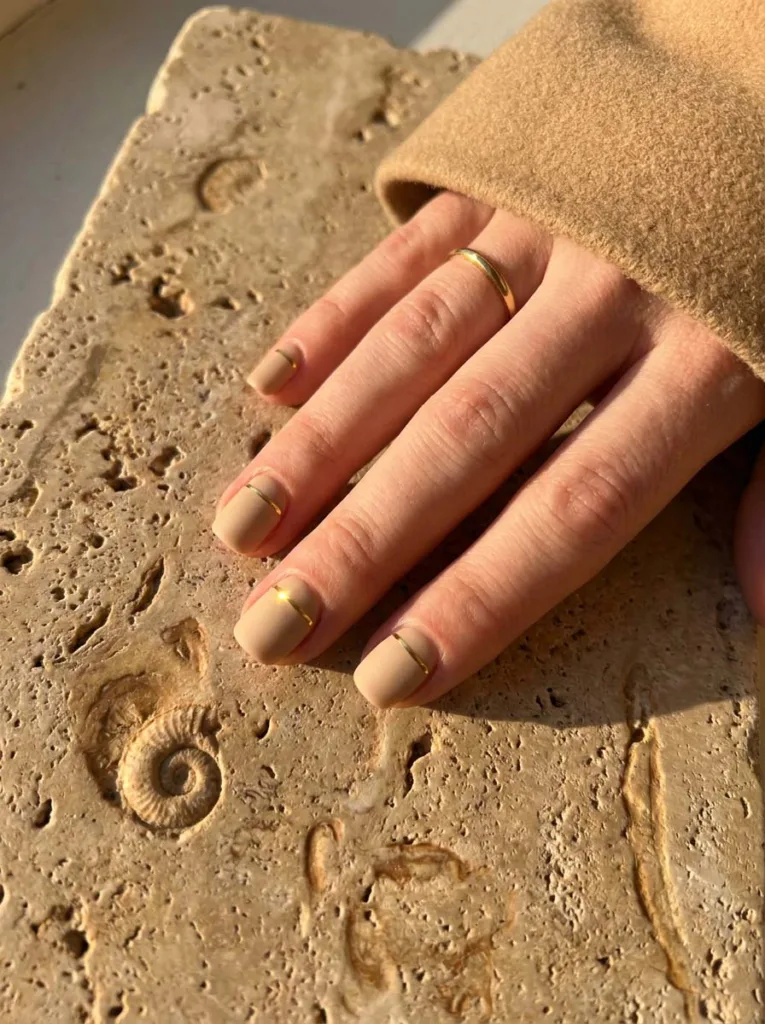 Warm beige nail polish with a single thin horizontal gold line near the cuticle on short squoval nails