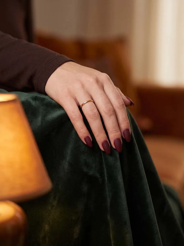 deep burgundy matte velvet almond nails on fair skin resting on dark green velvet fabric