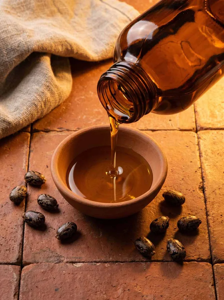 thick golden castor oil poured into ceramic bowl with castor beans on rustic terracotta tile surface