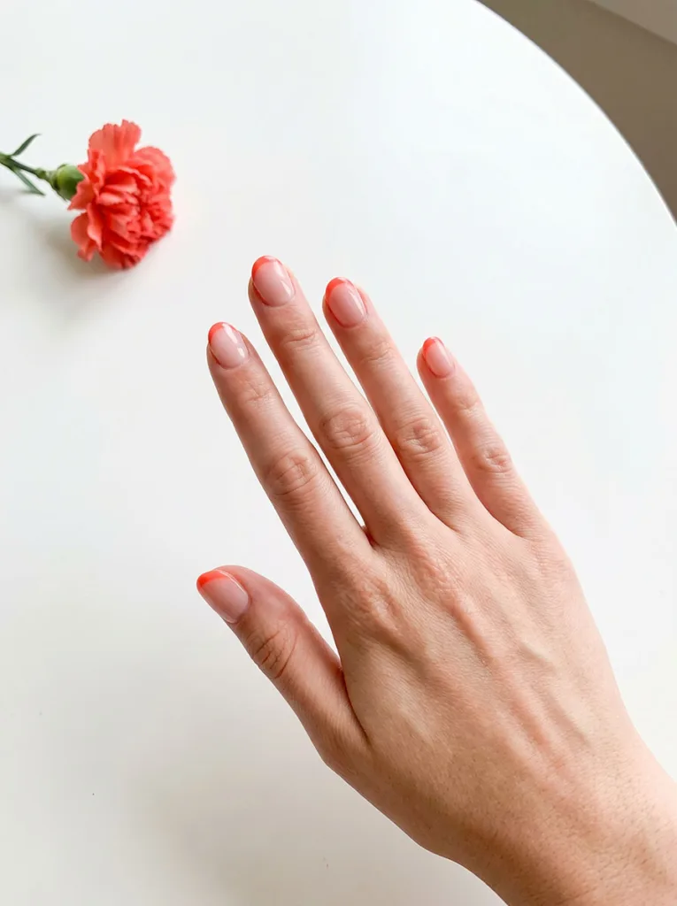 Coral French tip on sheer base, short round nails on fair skin with coral carnation accent prop