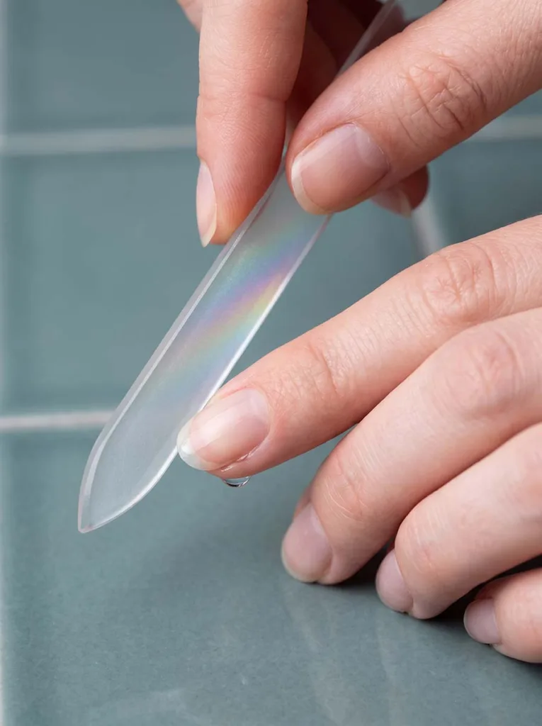 close-up of a glass nail file being used in one direction on a natural oval nail, fair-skinned hand