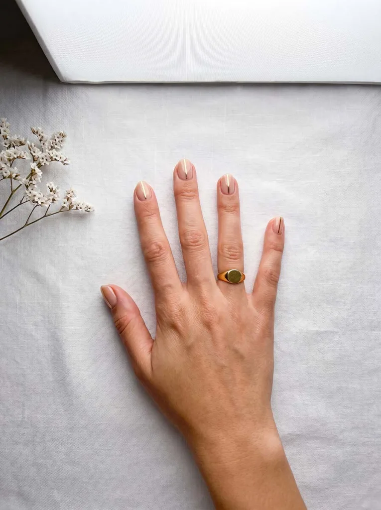 Gold vertical line nail art on short round nails, fair skin, overhead editorial on white linen