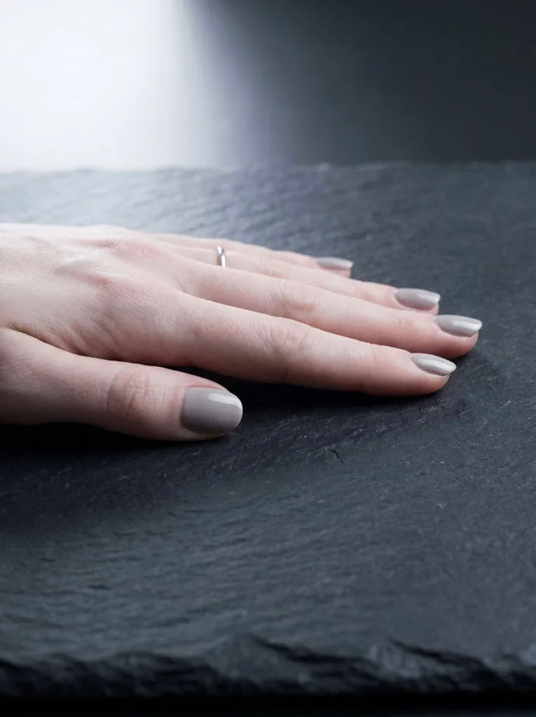 Greige short round nails on very fair skin in side-profile view on dark charcoal slate — modern editorial