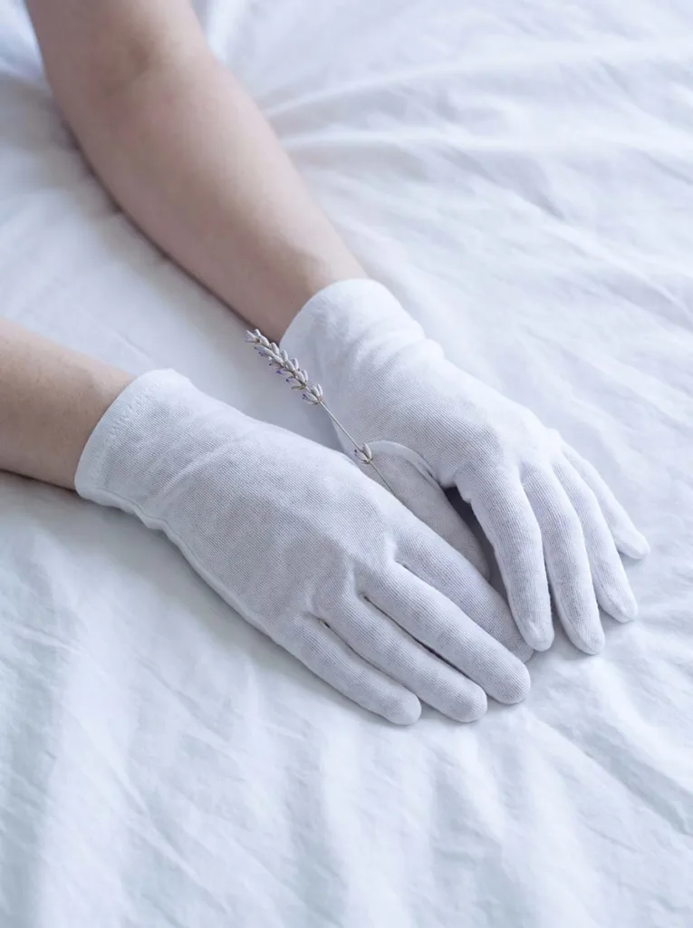 fair-skinned hands wearing white cotton gloves resting on white bed sheets for overnight cuticle oil treatment