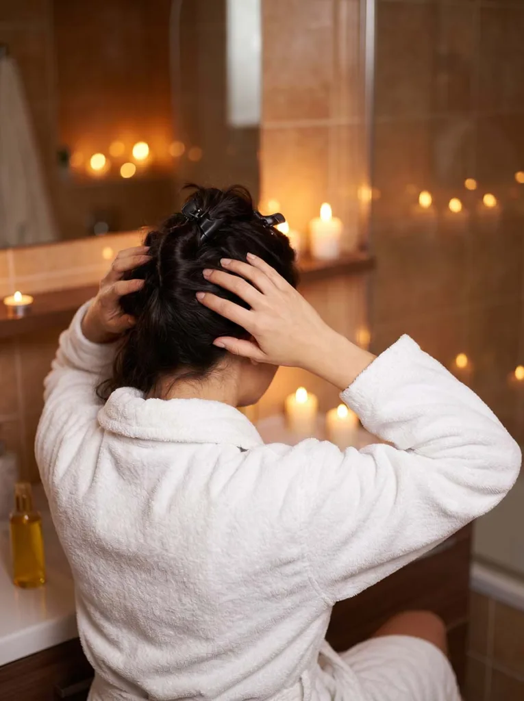 woman in white robe applying hair oil to scalp with fingertip massage in candlelit bathroom