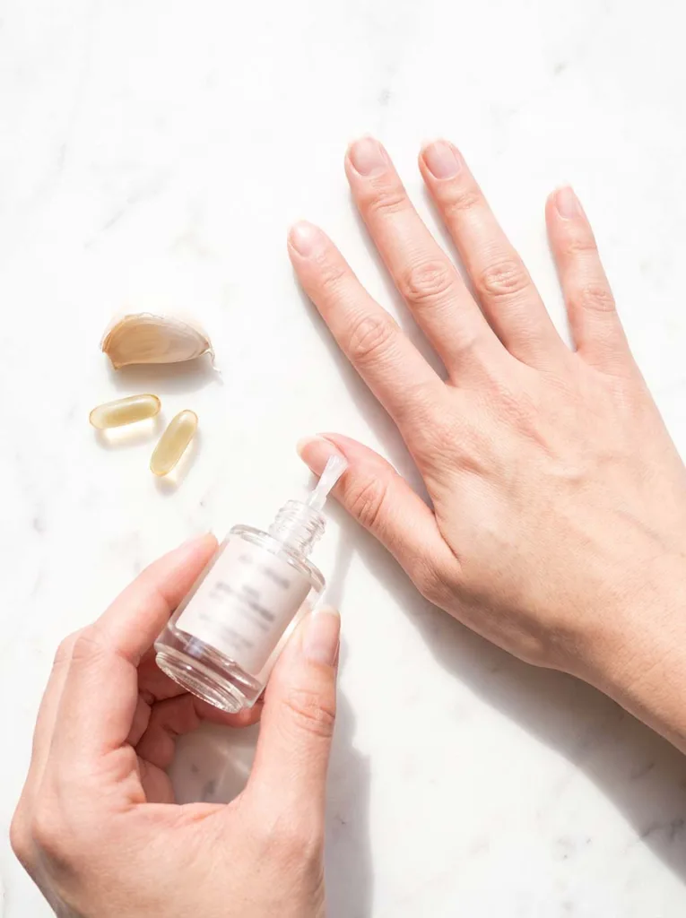 woman applying a clear nail strengthener during her monthly polish break week, bare nails on white marble surface