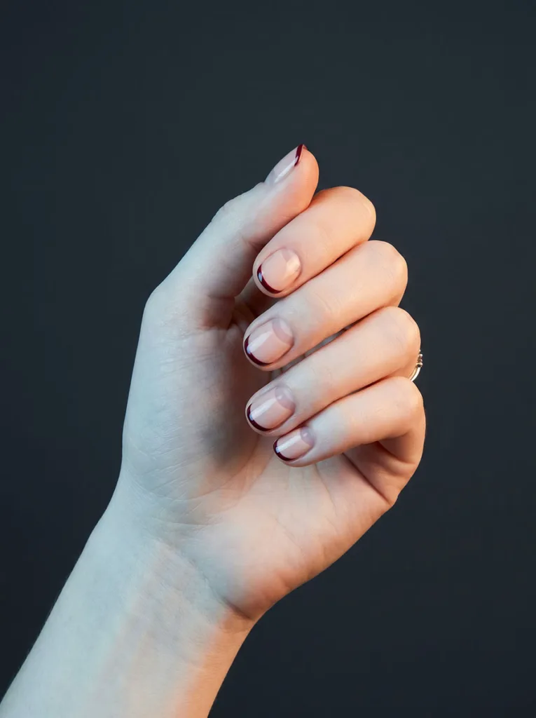 Negative space French short round nails on fair skin — deep burgundy edge line on nude base against charcoal backdrop