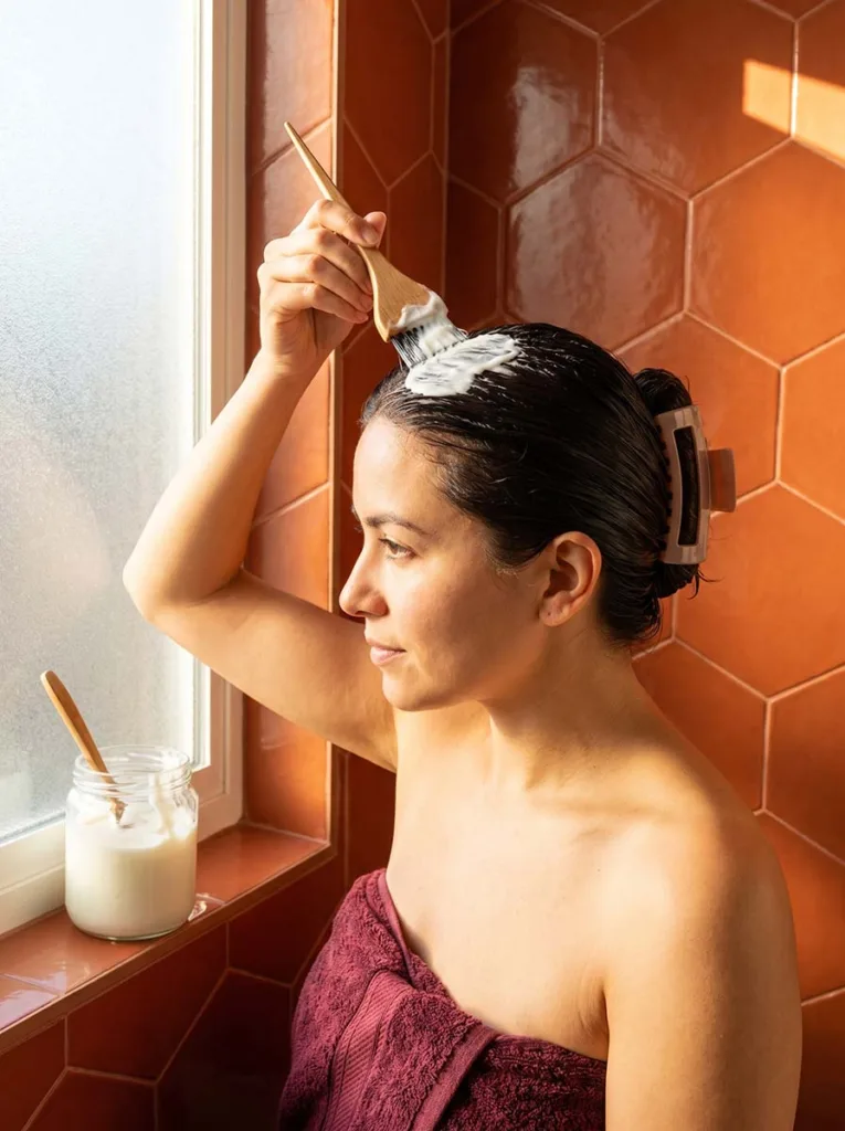 oman with wet hair applying plain white yogurt to her scalp with a small wooden brush while sitting in a bright bathroom with terracotta tiles