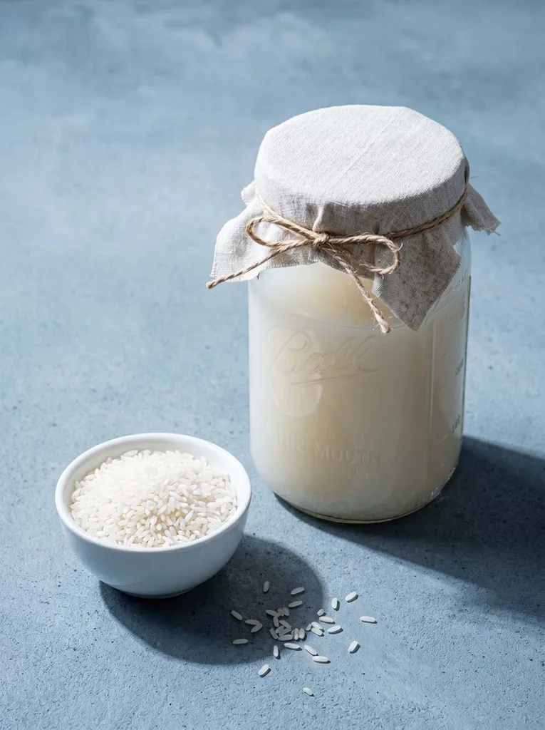 Glass mason jar of cloudy fermented rice water beside a bowl of uncooked rice on a blue-grey cement surface