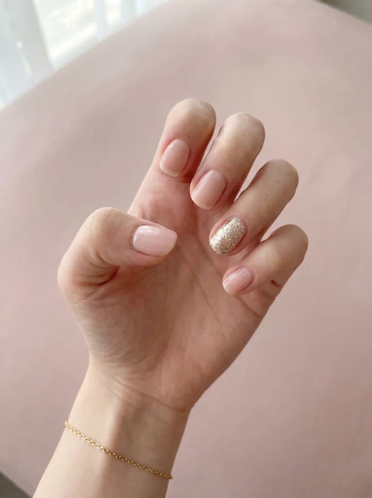 Sheer pink short round nails on fair skin with champagne glitter accent ring finger — soft-light editorial