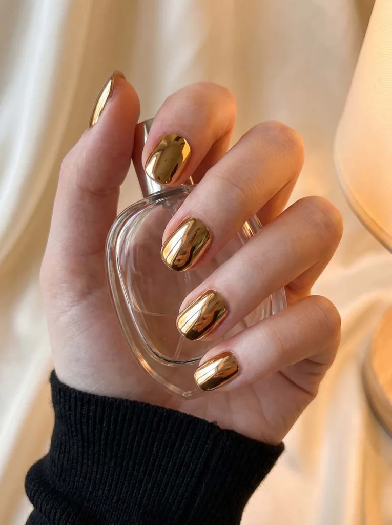 Soft gold chrome short round nails on fair porcelain skin holding a clear perfume bottle — warm-light editorial