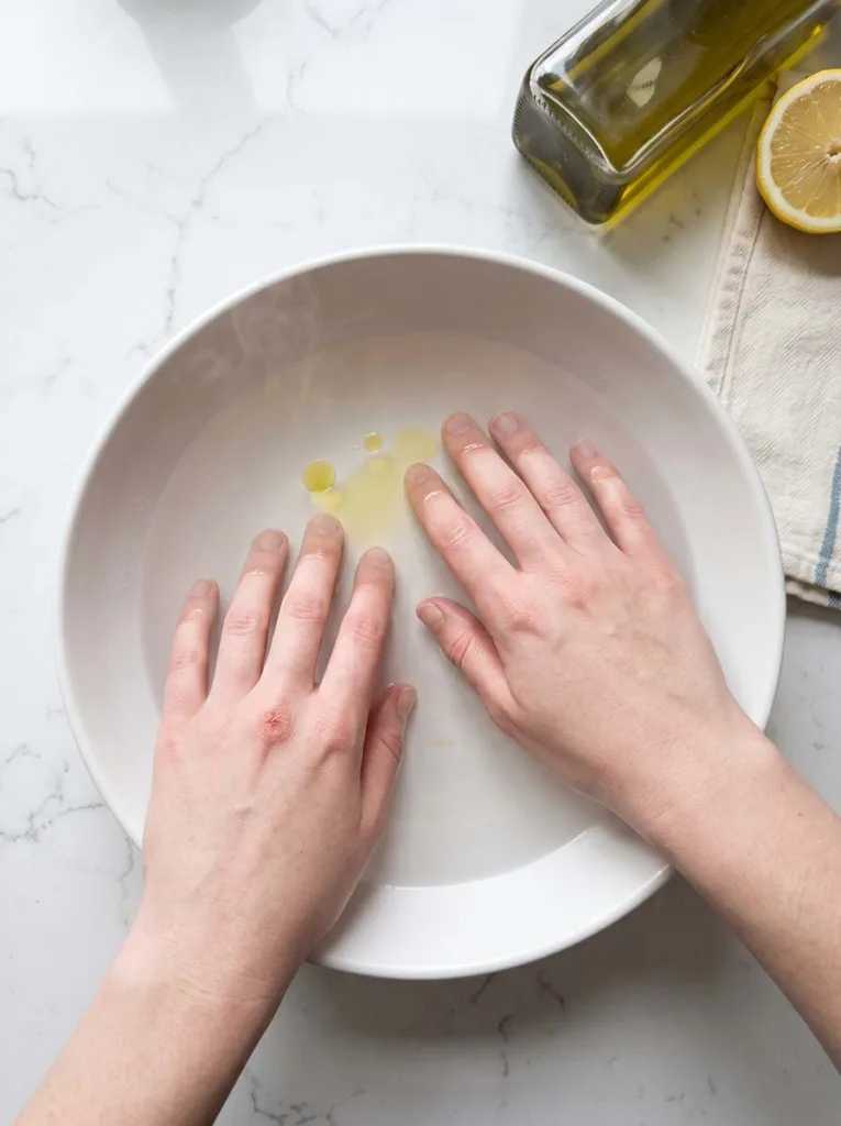 Hands soaking fingertips in a bowl of warm water with olive oil drops on a kitchen counter for at-home nail care