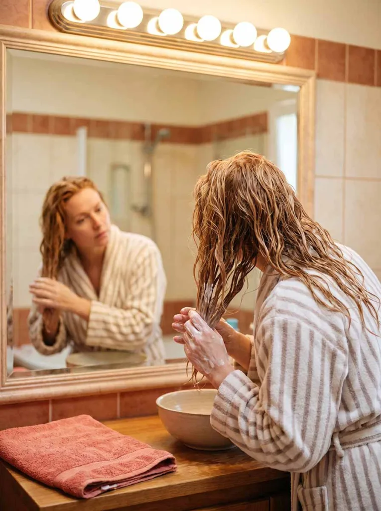 woman applying hair conditioner from mid-shaft to ends while standing at a bathroom mirror