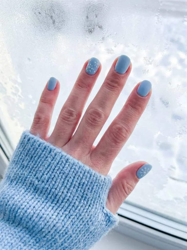 Baby blue winter nails with white snowflake accents near frosted window