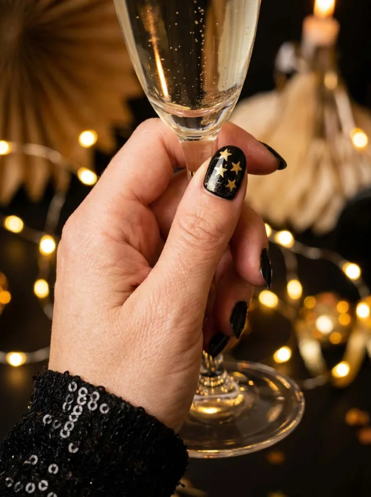 Black Christmas nails with gold star accents beside a champagne flute