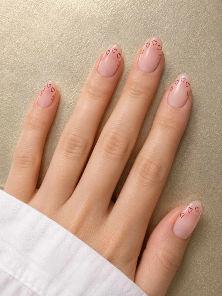 Almond nails with tiny red heart outlines replacing the French tip in a loose arc across the nail tip on a gold metallic background