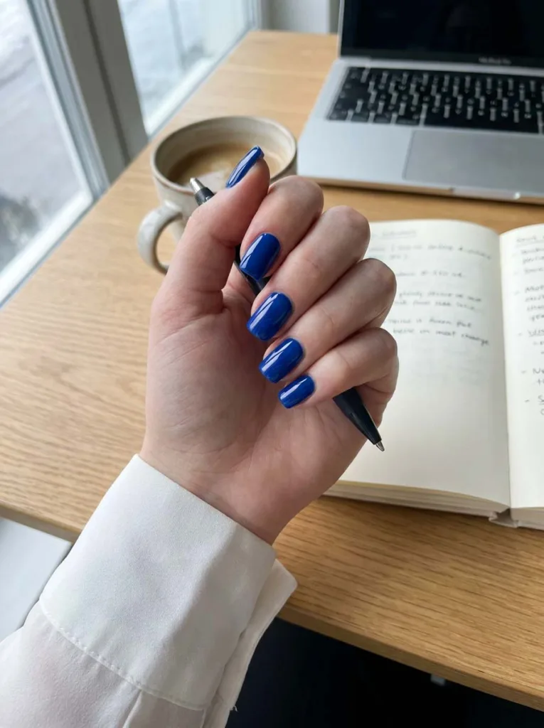 hand with glossy royal blue nails resting on a wooden desk near a coffee cup