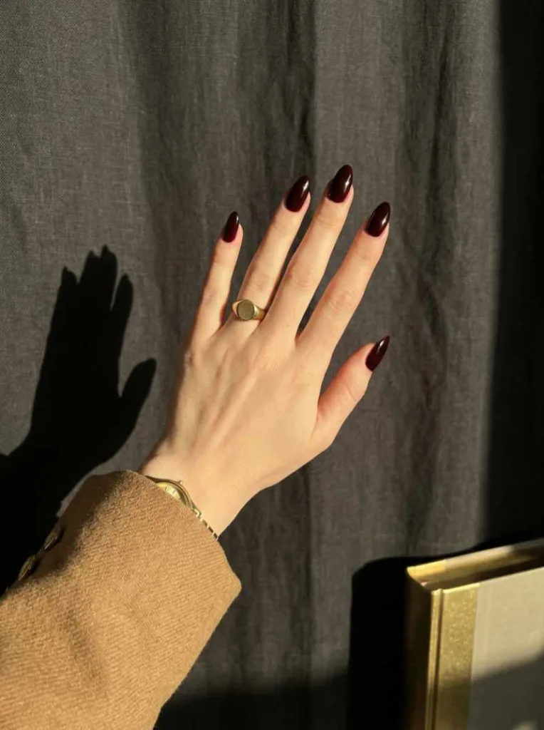 Almond-shaped deep burgundy nail polish with creme finish on fair hand against dark charcoal fabric with gold blazer cuff