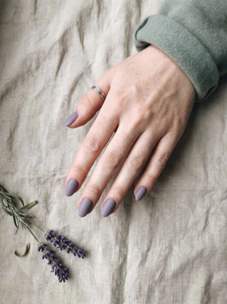 Fair hand with dusty lavender matte nail polish and silver ring resting on natural beige linen fabric with dried lavender