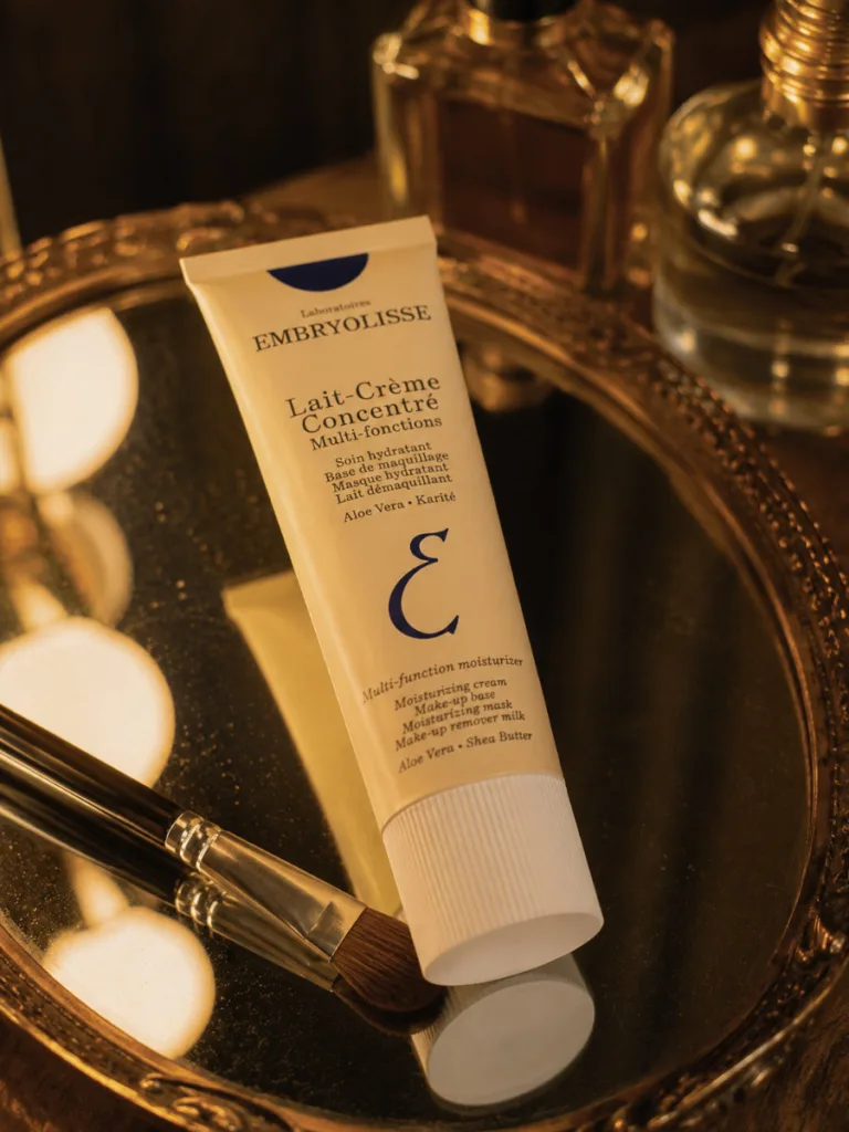 Embryolisse Lait-Crème Concentré tube resting on a mirrored vanity tray beside a makeup brush