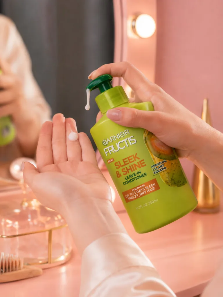 Hand applying Garnier Fructis Sleek and Shine leave-in conditioner at a vanity table