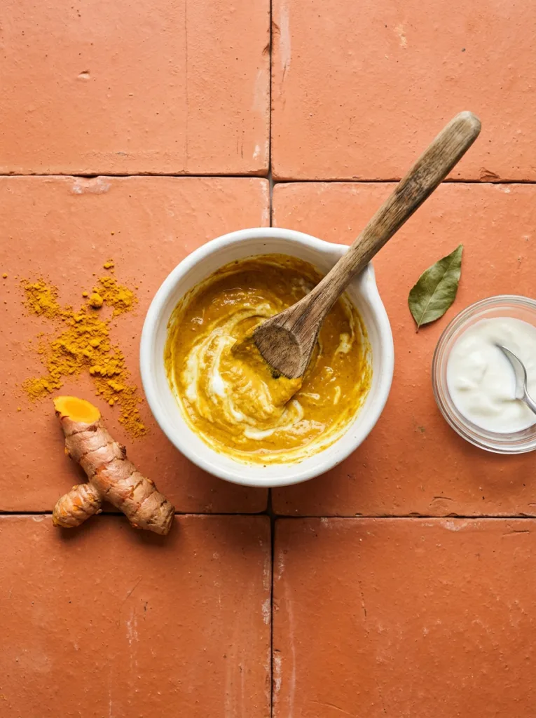 Golden turmeric and yogurt DIY face mask being mixed in a small white ceramic bowl with a wooden spoon on a terracotta tile surface