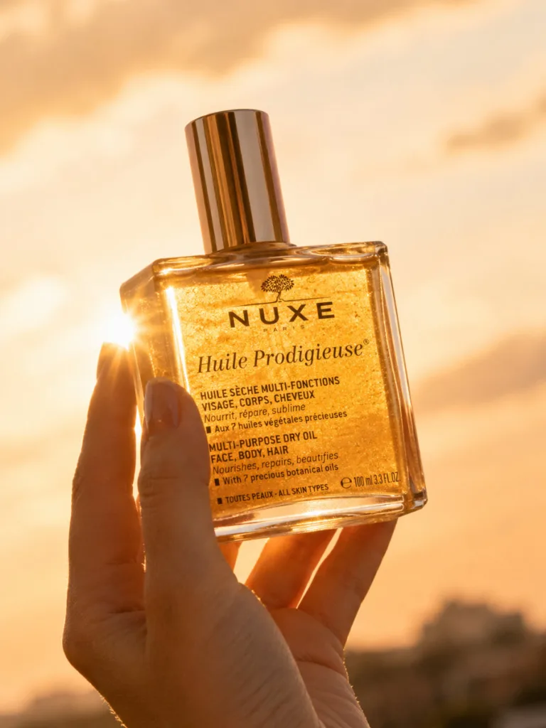 Nuxe Huile Prodigieuse gold-tinted dry oil bottle held up against golden sunset light