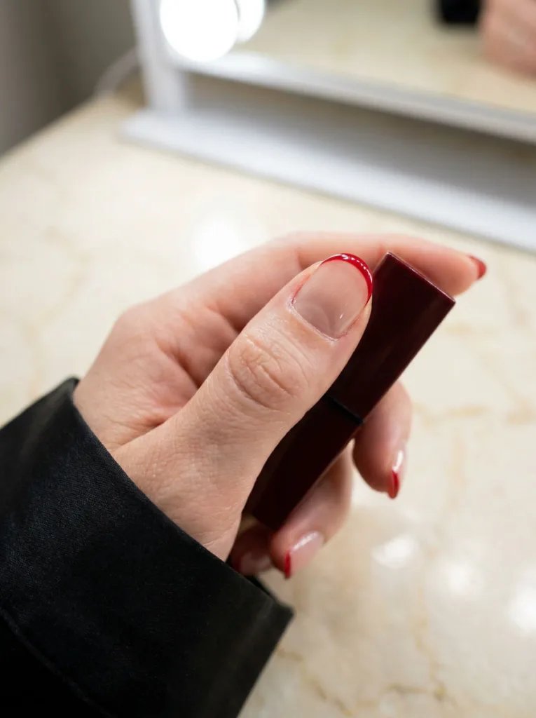 Micro french tip red christmas nails on a hand holding lipstick at a vanity